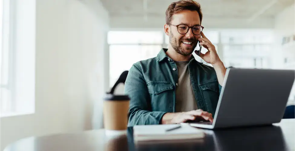 Smiling man with beard and jeans jacket talking on the phone while looking at a laptop on his desktop