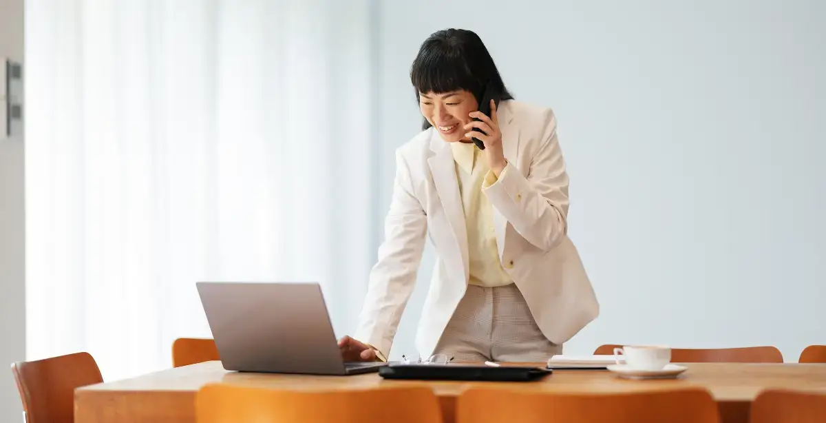 Business woman in office setting talking on the phone while typing on a laptop.